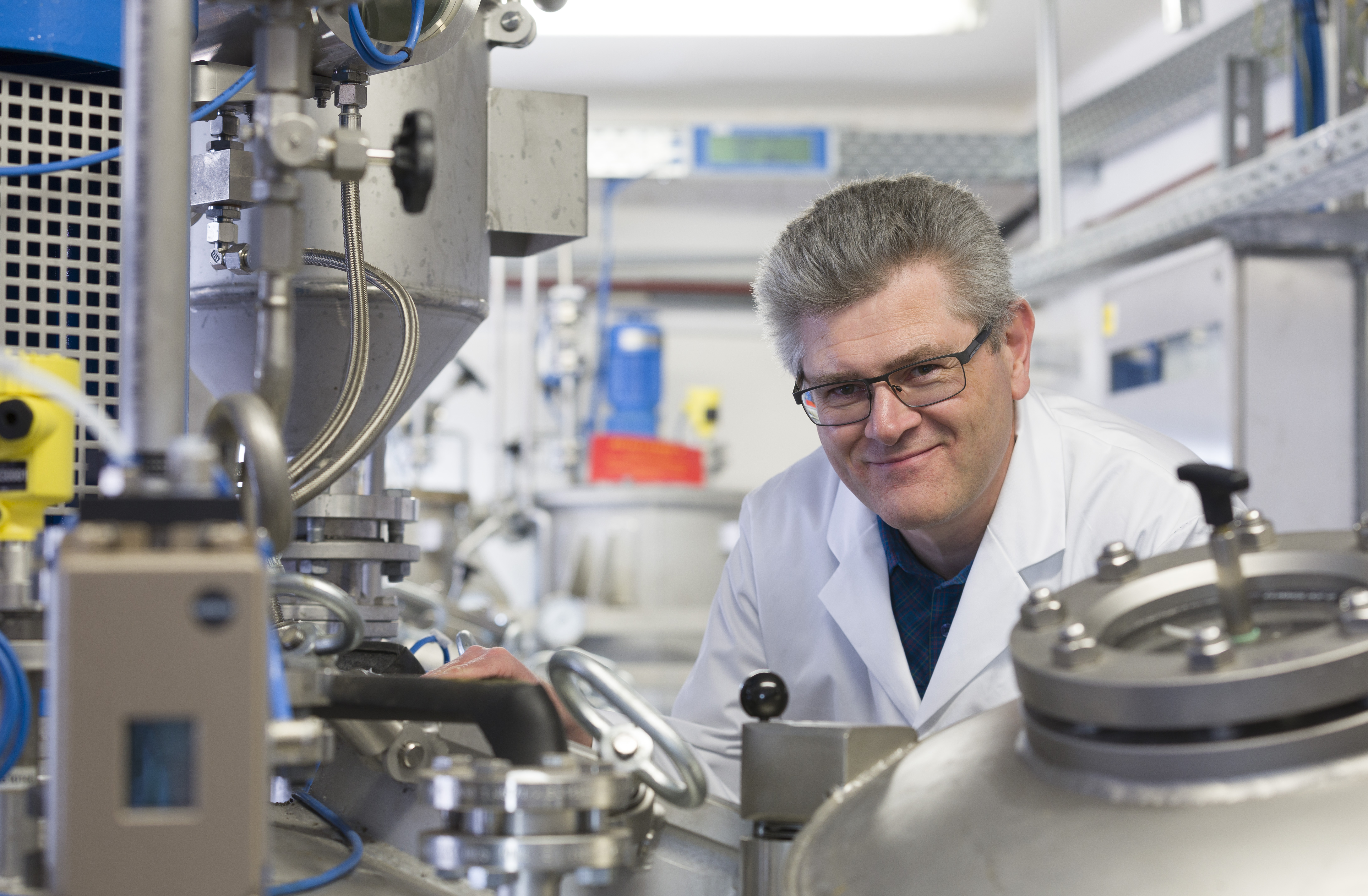 Dr. Markus Lettau im weißen Laborkittel und mit Brille lächelt, während er in einer wissenschaftlichen Einrichtung oder Forschungseinrichtung zwischen großen industriellen Laborgeräten und Maschinen arbeitet.
