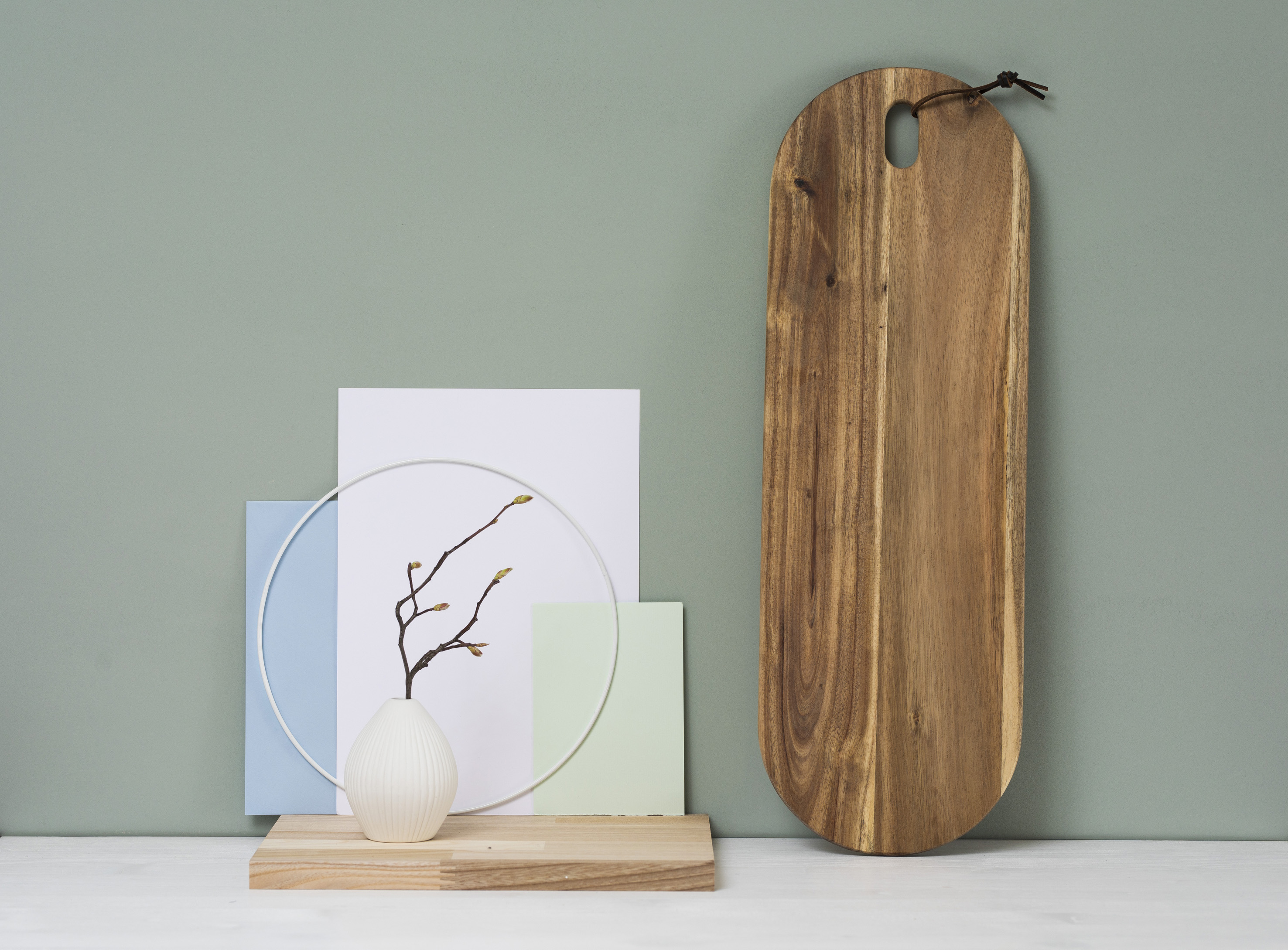 Ein minimalistisches Arrangement mit einer kleinen weißen Vase mit einem Zweig, pastellfarbenen Karten und einem runden Draht auf einem Holzsockel, neben einem hohen Holzschneidebrett vor einem gedämpften grünen Hintergrund.