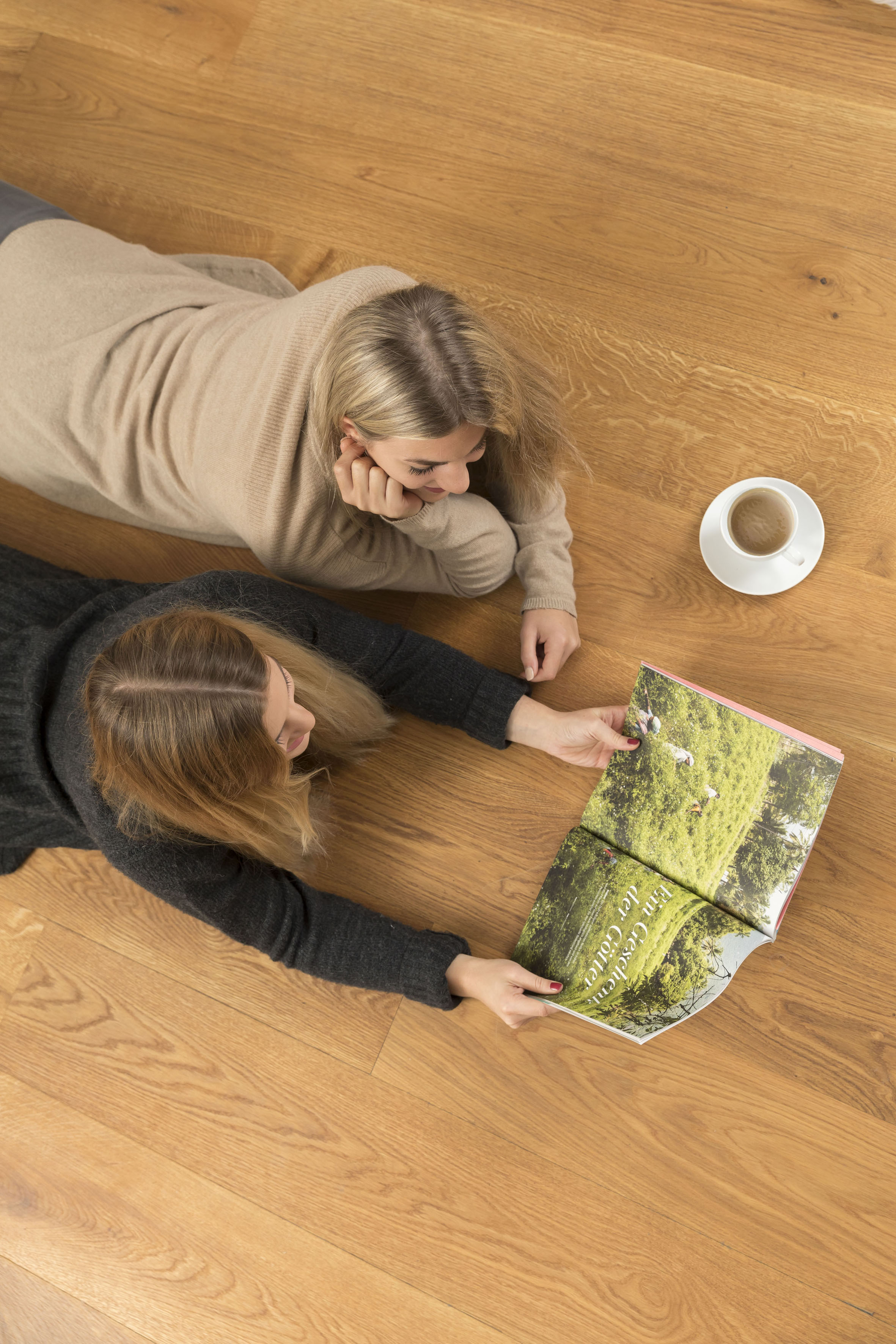 Zwei Frauen liegen auf einem Holzboden und betrachten eine aufgeschlagene Zeitschrift mit Naturfotos. Daneben steht eine Tasse Kaffee auf einer weißen Untertasse.