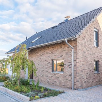 Ein modernes Backsteinhaus mit dunklem Ziegeldach, mehreren Fenstern und einem kleinen, grünen Garten davor. Der Himmel ist teilweise bewölkt, und das Haus ist von einem gepflasterten Gehweg umgeben.