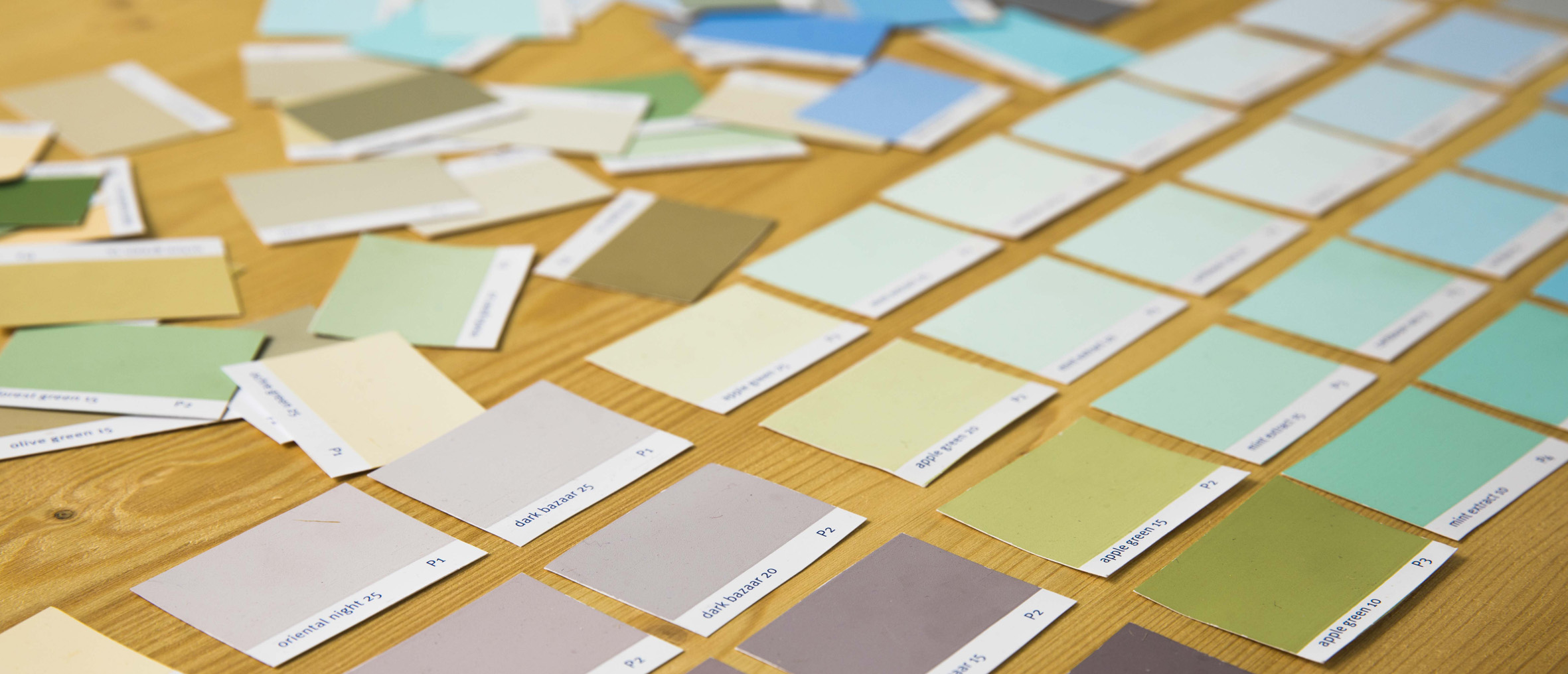 Mehrere Farbmuster in verschiedenen Grün-, Blau-, Beige- und Grautönen liegen geordnet auf einem Holztisch. Manche Muster sind ordentlich in Reihen angeordnet, andere liegen verstreut herum.