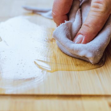 Eine Hand trägt mit einem Tuch Öl auf ein Schneidebrett aus Holz auf und bereitet so die Oberfläche für die Wartung und Pflege vor.