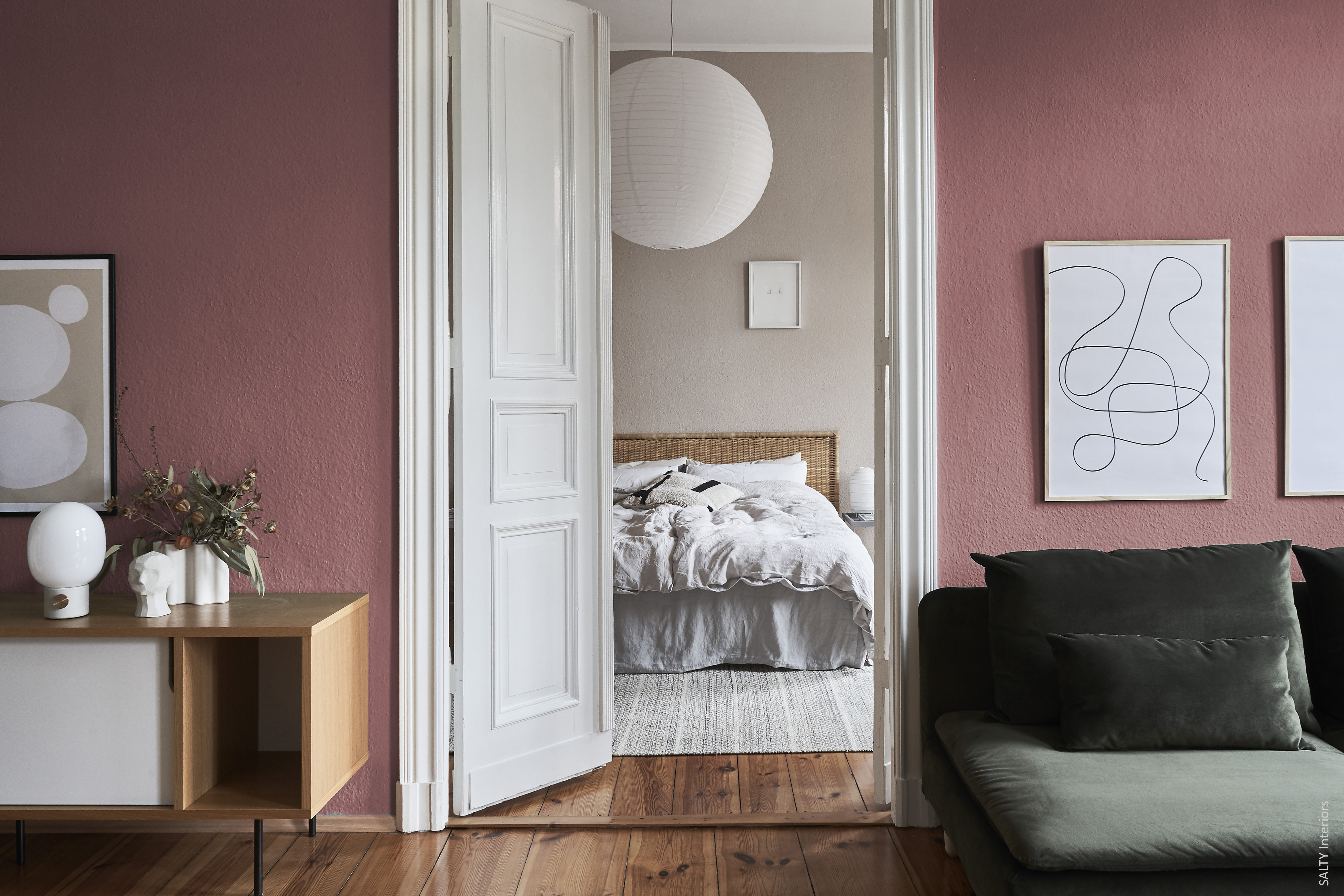 Helles Wohnzimmer in Rosa mit grünem Sofa, offener Tür zum Schlafzimmer mit Korb-Kopfteil und weißer Bettwäsche.