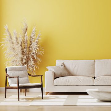 Ein modernes Wohnzimmer mit einer gelben Wand, einem beigen Sofa, einem passenden Polstersessel, einem hellen Teppich, einem weißen Couchtisch und einem hohen Pampasgras-Arrangement in der Ecke. Sonnenlicht erhellt den Raum.